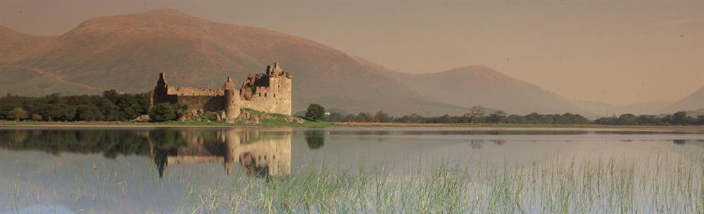 Picturesque ruins of Kilchurn Castle with mountains and sky in background, shown reflected in still water