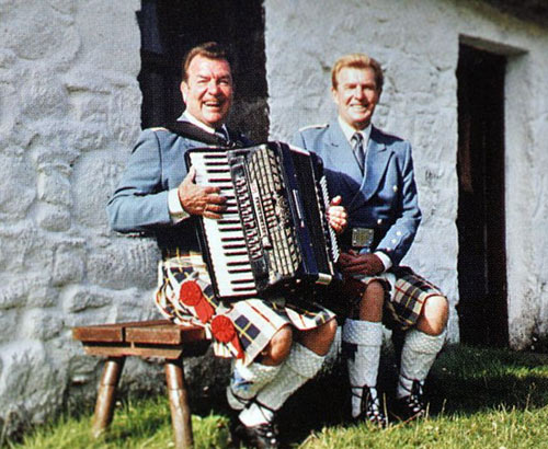 The Alexander Brothers seated in front of a white cottage wearing blue jackets and black, red and yellow kilts, one holding an accordion