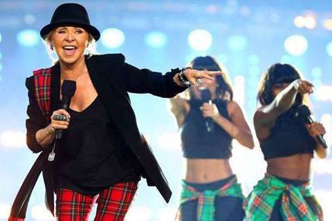 Lulu wearing black hat, tartan trousers and black suit jacket singing with two female backing singers